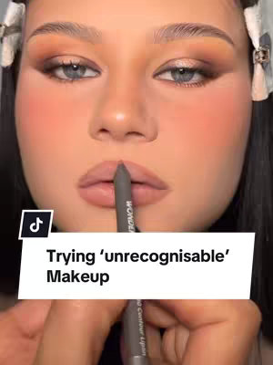 A glamour shot featuring a woman's face in close-up, with soft lighting enhancing her features. Her skin has a smooth texture with a blush effect. Her eye makeup is a blend of browns. She holds a lip contouring pencil, poised over her lips, with a text overlay near the bottom of the frame.