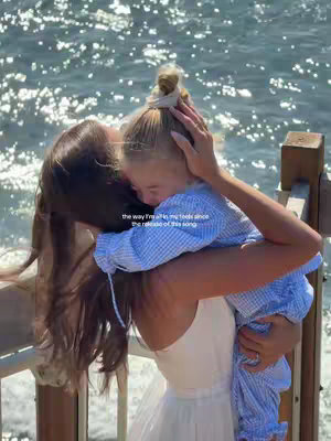 A woman is holding a young child. The background is out of focus, with the ocean and a wooden structure visible. The woman has dark hair and is wearing a white dress, hugging a child with blonde hair, wearing a light blue outfit. She appears to be embracing the child tightly. The lighting suggests it's daytime.