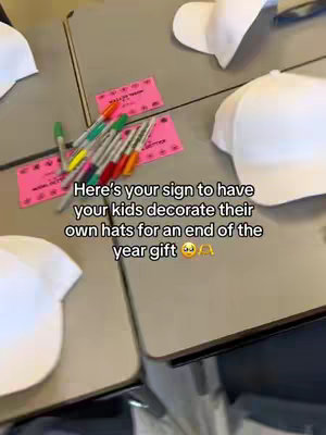 A top-down shot of a light-colored table with several white baseball caps, pink paper cards, and colored markers. The text overlay reads, "Here's your sign to have your kids decorate their own hats for an end of the year gift".