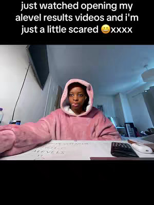 A young woman in a pink hoodie is seated in front of a desk. The background shows a room with a window, a partial view of a television, and various home items. The text overlay at the top reads: "just watched opening my alevel results videos and I'm just a little scared xxxx". The woman has a slightly distressed expression, which includes her tongue sticking out and her eyebrows furrowed slightly.