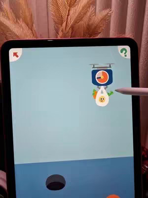 An iPad displaying a game's interface is the central focus. The game screen showcases a minimalist design with colorful, cartoonish elements like fruits, weighing scales, and cooking-related icons. The iPad is held upright. A white stylus is positioned, suggesting user interaction. The background shows neutral-toned fabric and floral decorative elements.
