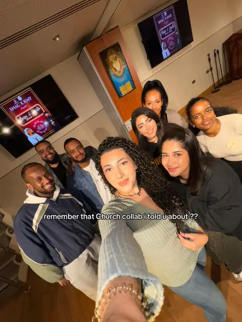 A selfie-style photo featuring a group of eight people, predominantly women, indoors. The group is closely arranged, with the primary focus on the person holding the camera, a woman with curly dark hair and a light green top, positioned in the center. She's surrounded by the others, with several women to the right, and men to the left. Behind the group, there are two screens displaying what seems to be promotional materials, with text and graphics related to an event titled 'SNAC X KMY'. The background includes neutral-toned walls and some decorative elements. The overall lighting is warm, and the composition centers on the group's faces for a friendly, inviting feel.