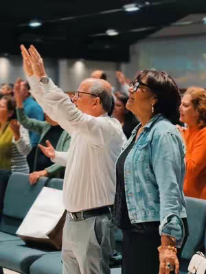 An interior shot of a church. Two adults are the main focus, framed from the waist up, standing near the middle with their arms raised. A man on the left wears a white shirt and gray pants; a woman on the right wears a denim jacket over a black top. They are surrounded by a congregation with raised hands. Rows of pews and a large screen are visible in the background. The lighting is soft, creating a sense of reverence, and the overall palette is muted.