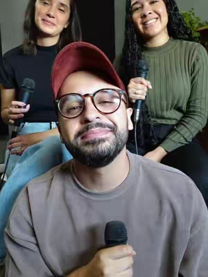 A cover photo showing a man front and center holding a microphone and smiling. Two women are positioned behind him, each holding a microphone. The man is wearing glasses and a maroon cap. The women are smiling in the background.