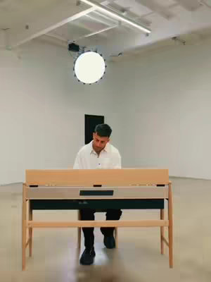 A man is seated at a light-colored wooden piano, playing. The background is a minimalist white space, with a bright circular light fixture overhead and a dark vertical element behind the man. The lighting in the room is bright and evenly distributed.