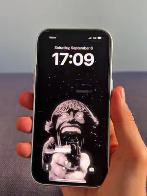 A hand holds an iPhone displaying a black and white live wallpaper of a person with a gun. The phone screen also shows the time and date.