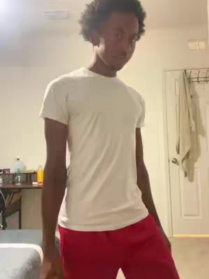 A person of color wearing a white t-shirt and red shorts stands indoors. The setting is a room with beige walls, a white door in the back, and a bed partially visible on the left. The person has short, curly hair and is looking slightly downward. The lighting is ambient.