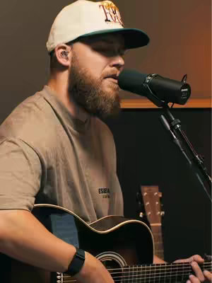 A man with a beard wearing a baseball cap and a t-shirt, is focused on singing into a microphone while playing a guitar. The background is dark, with studio equipment visible.