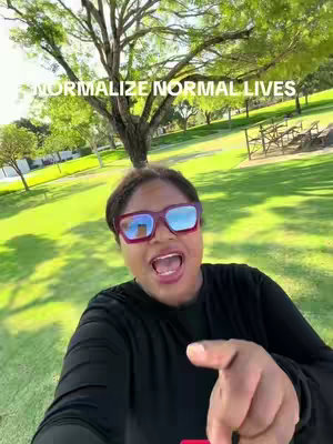 A selfie-style shot of a woman in sunglasses, smiling with her mouth open wide, pointing her right finger toward the viewer. She’s in a park setting with green grass, trees, and the sky visible in the background. The text overlay indicates that this content revolves around normalizing normal life.