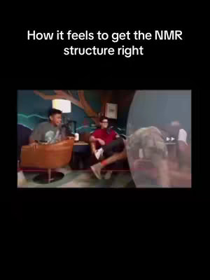 The image shows a scene with three people in a well-lit interior setting, likely a studio or office. Two men are seated in the foreground; one is in a grey sweatshirt and seated to the left, appearing to speak while leaning forward with a microphone, the other in a red shirt is seated on the right. Behind them, a third person, partially visible, is seemingly reacting to something with a surprised expression; this image shows a video overlay of transparent layer with an expressive reaction and text hook.