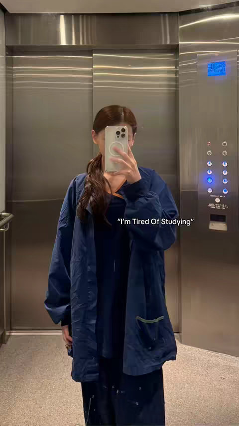 A woman is shown in front of a metallic elevator. She is holding a phone in front of her face, obscuring her features. She is wearing a navy blue jacket and the photo has cool lighting. A text overlay displays "I'm Tired Of Studying".