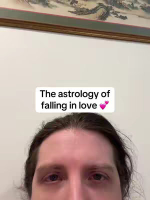 The cover image depicts a person with long hair from the forehead down, centered in frame, with a white box overlay at the top, containing the text "The astrology of falling in love" in a simple sans-serif font. There are two pink heart emojis next to the text. Behind the person the backdrop is a white wall, with the edge of a picture frame visible.