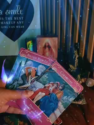 The image shows a tarot card reading setup. The foreground features a hand holding several tarot cards, arranged to display their images. Behind the hand, there's a dark background, with various items. The lighting is dim, casting a mysterious and spiritual ambiance.