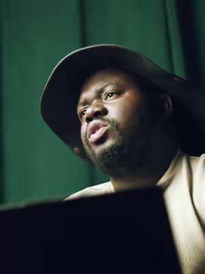 A man wearing a hat speaks with his mouth open, lit by a green background.