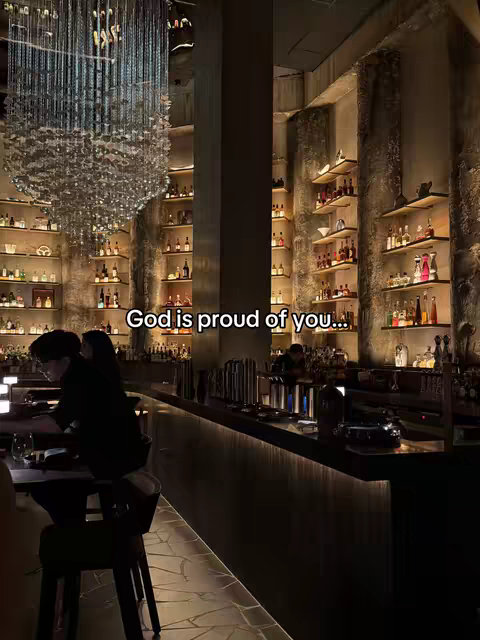 An interior shot of a dimly lit bar with shelves stocked with bottles, a decorative chandelier, and a person sitting at the bar in the foreground. The text overlay on the image reads 'God is proud of you...'.