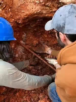 Two people wearing hard hats and gloves are reaching into a brown cave-like opening with a tool. The opening has rough textures, and they appear to be working together to remove an item, possibly a crystal.