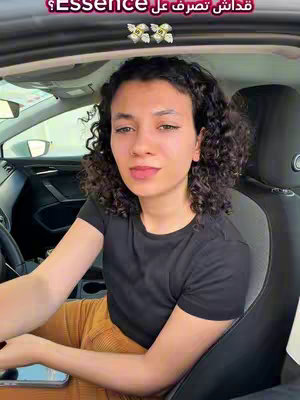 A woman with curly hair sits in the driver's seat of a car, facing the camera. She is wearing a black t-shirt and orange pants. Text in Arabic is overlaid at the top of the image.