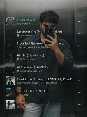 A selfie taken in an elevator, featuring a person holding a phone in front of their face. The elevator walls are mirrored, partially reflecting the person. A Spotify playlist appears on the left side of the image, showing song titles and artist names. The person is casually dressed in a dark button-down shirt and light-colored pants.