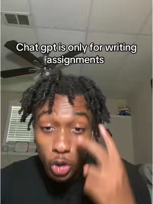 A young Black man appears close-up, looking directly at the camera with a slight frown. He has short, twisted black hair and is wearing a black shirt. Above him, a text overlay reads, 'Chat gpt is only for writing assignments.' His expression suggests a mix of acknowledgment and a hint of amusement. The background is blurred, featuring a ceiling fan and a section of a white wall. The overall composition is simple and direct, focusing on the man and the text.