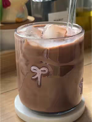 The image shows a clear glass filled with a thick, brown liquid, likely a chocolate beverage, with ice cubes floating on top. A glass straw is inserted into the drink, and the glass rests on a white coaster. The background is blurred, suggesting the focus is on the drink.