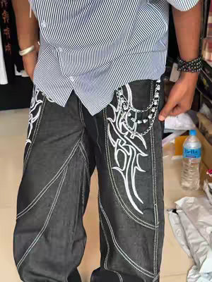 A person is shown from the waist up, wearing a striped button-down shirt and black denim jeans with intricate white tribal-style embroidery. A silver chain with charms hangs from the jeans' pocket. Other items in the background like water bottle are slightly visible.