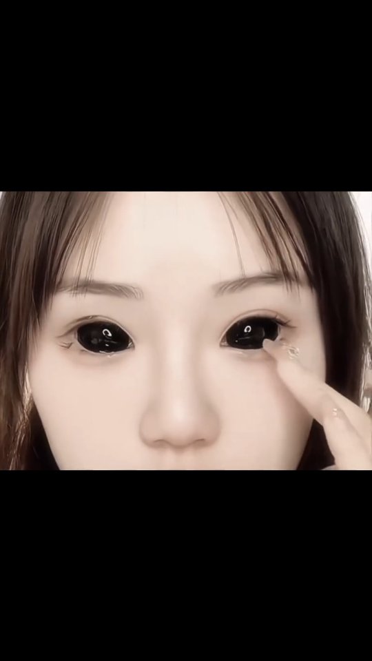 A close-up shot of a person's face, focusing on their eyes, which are entirely black. They are touching their face with a finger. Their skin is pale with minimal makeup and the upper portion of the body is visible. The background is neutral