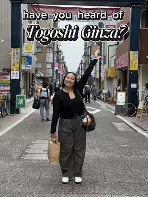 A woman stands in a street in Japan, smiling and raising her arm in a welcoming gesture. The background shows a shopping street with signs and shops.