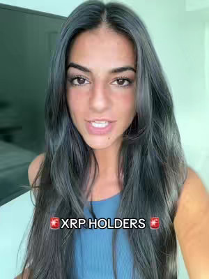 A woman with long, dark hair is centered in the frame, looking directly at the camera and speaking. Overlayed on the video near the bottom, in a box, is the text “XRP HOLDERS”. The background appears to be a room with a gray door or wall and a glimpse of a bed.