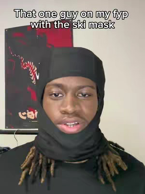 A man with dreadlocks wearing a black ski mask is centered in the frame. Behind him, a poster with a red and black design is visible. The background is a plain white wall.