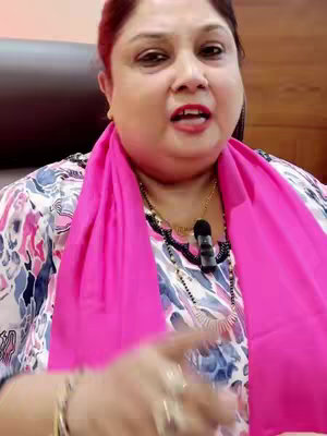 A medium shot features a woman with fair skin, dark hair, and a bright pink scarf. She is positioned slightly off-center, looking towards the viewer with a warm expression. Her outfit includes a patterned top with a white and blue floral design. The background appears to be a muted interior space, suggesting a home or office setting. The woman's hand is in the frame, and she also wears jewelry including a neck chain and rings.