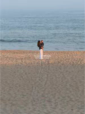 A woman stands on a sandy beach holding a young child, facing the ocean. The background consists of the sea and sky, with text overlayed at the bottom.
