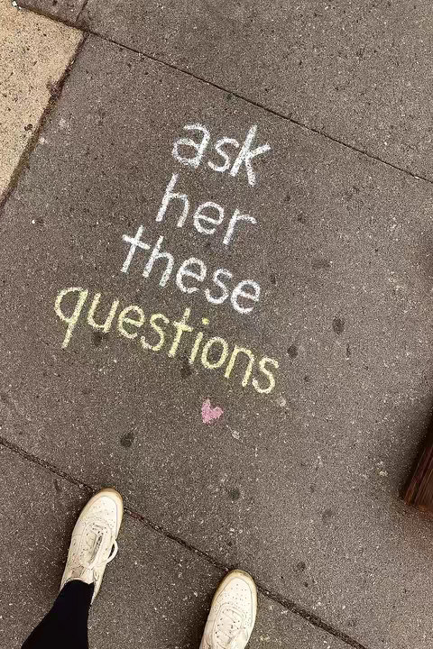 The image shows a close-up of a gray pavement. Chalk-written text in white and yellow is in the center, reading "ask her these questions" with a small pink heart. The bottom of the image shows two white sneakers. The colors are muted, with a focus on the texture of the pavement and the handwriting style of the chalk.