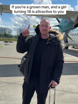 A man in a black jacket and black pants stands on an airfield, pointing his finger upwards. A plane is visible in the background with the sky above.