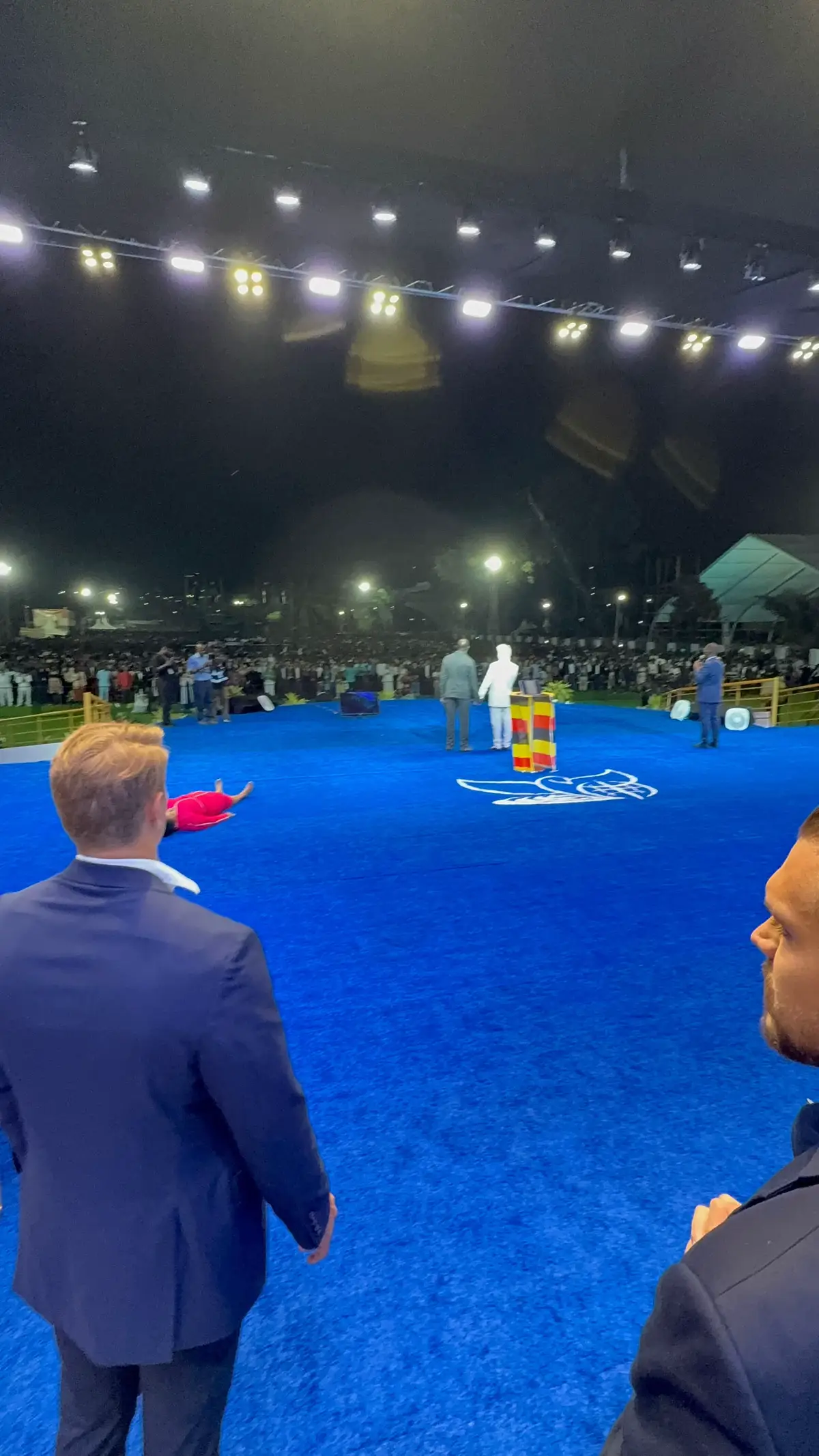 The cover image shows an outdoor event at night. There's a large blue rug or stage, and two men in suits stand in the center, facing each other near a small, colorful podium. Behind them is a large crowd, and stage lights are in the background, adding to the ambiance.