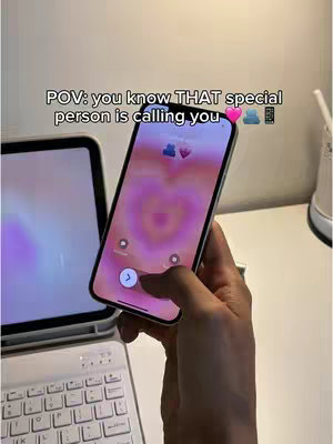 The image shows a hand holding a phone with a pink and purple heart-shaped background, suggesting a call from a special person. A laptop and keyboard sit in the background.