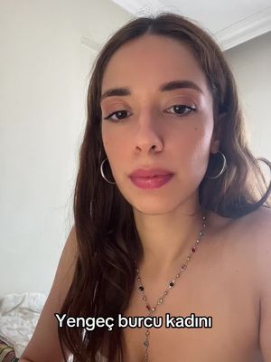 A medium shot features a young woman with long brown hair and light skin looking directly at the camera. She has a neutral expression, with her eyes looking forward and subtly defined lips. She has a necklace of colorful stones. The background is a simple, off-white wall. Text at the bottom of the image reads, 'Yengec burcu kadini'.
