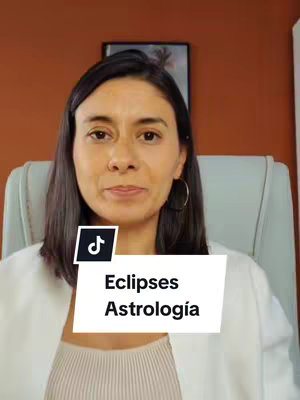 A woman with dark hair and a white blazer sits in an office chair. A text overlay says 'Eclipses Astrologia'. Behind her is an orange wall and a picture frame.
