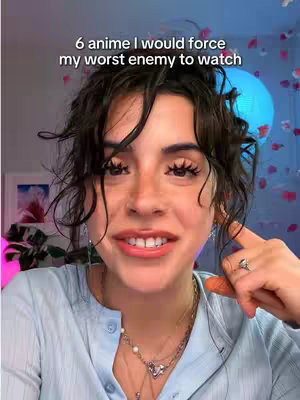 A close-up shot of a woman with dark curly hair and fair skin, wearing a light blue shirt and several necklaces. She is smiling slightly and looking directly at the camera. The background is softly blurred with hints of pink and blue lighting, and some decorative elements that appear to be jellyfish or flowers.