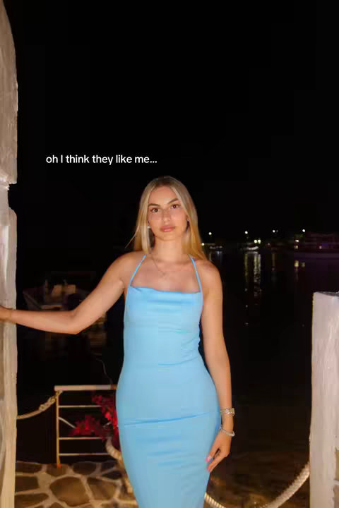 The cover image depicts a young woman standing outdoors at night. She is wearing a light blue, fitted dress. The background features a body of water with lights reflecting on it, suggesting a bay or harbor. The woman is positioned near a white structure, possibly a pergola or building detail, upon which she leans. The sky is dark but clear, and there are some lights in the distance. A text overlay is present, which reads, "oh I think they like me..."