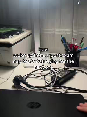A low-angle shot captures a desk workspace, with a laptop, power adapter, and pens in clear view. Text overlays read, indicating a study or work-related scenario. The setting suggests a home or office environment.
