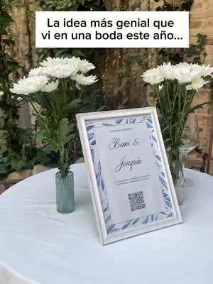 The cover image shows a table set for a wedding. On the table is a white framed sign with a printed invitation and a QR code, and two vases with white flowers and greenery. The background hints at a lush outdoor setting.