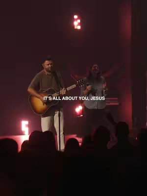 Two men, one playing an acoustic guitar and singing, the other singing with his hands in the air, are performing on a stage. Text overlayed reads 'IT'S ALL ABOUT YOU, JESUS'.