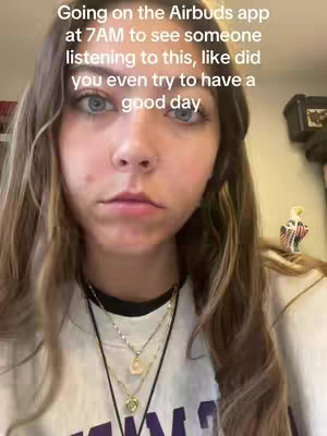 A selfie-style shot featuring a young woman with long, wavy brown hair. She has fair skin, with visible skin texture. Her face is centered, and she has slight eye bags and a neutral expression. She wears a necklace with multiple small chain necklaces. The background is blurred, but shows hints of an interior setting, which is not in focus. The text overlay reads:"Going on the Airbuds app at 7AM to see someone listening to this, like did you even try to have a good day".