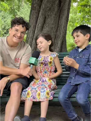 A medium shot shows three people on a wooden bench in front of a tree. A man with glasses sits to the left, holding a microphone towards a young girl in the center. A boy sits to the right, looking at the girl. The man on the left is wearing a light-colored t-shirt, and shorts. The girl is wearing a floral dress. The boy is wearing a denim jacket over a dark shirt and blue jeans. The background includes greenery, with blurred vegetation and a large tree trunk.