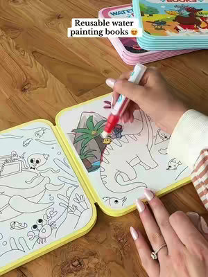 A person is holding a water pen and is painting a reusable water painting book. Several other similar painting books are on a wooden table. The book is open to a drawing, and the pen touches the drawing. The text "Reusable water painting books" is added as a hook.