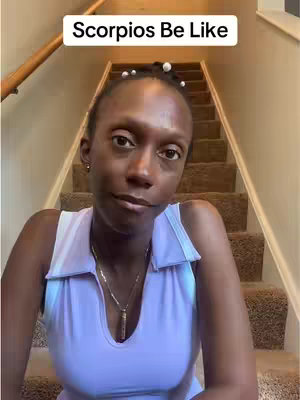A woman of color is seated on steps, looking toward the camera with a thoughtful expression. She is wearing a light-colored sleeveless top and a delicate necklace. The background shows a stairwell.