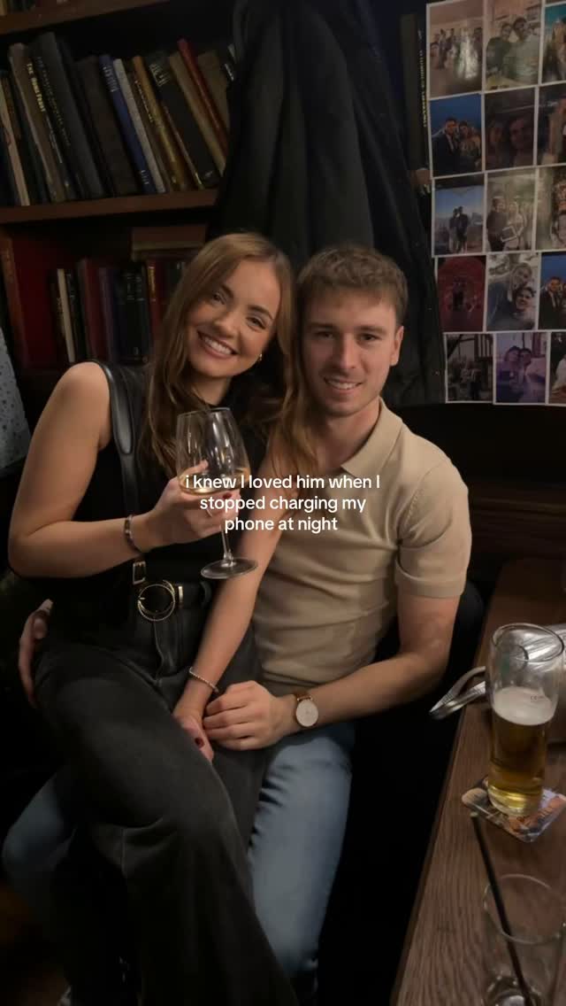 A couple sits close together in a dimly lit bar setting. The woman, holding a wine glass, smiles towards the camera. The man seated beside her is also smiling. The background includes a bookshelf and photos.