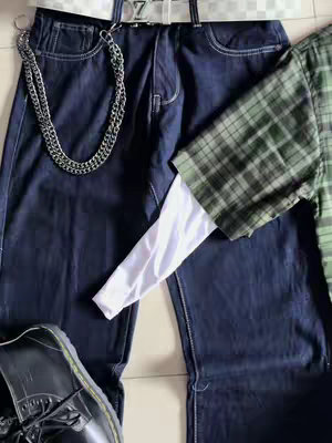 An overhead shot showcasing a set of layered clothing items and accessories. The dominant colors are dark navy for a pair of baggy jeans, and green and khaki green/brown, appearing in a plaid flannel shirt. A white long-sleeved shirt is layered underneath. The accessories include a silver chain attached to the jeans' belt loop, a silver belt with an embossed 'Z' buckle with a white belt, and a black shoe visible at the bottom left of the frame.