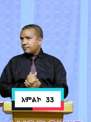 A man with a dark shirt and a purple tie is standing behind a podium. The cover image has a rectangular overlay with the text 'አምልኮ 33' on it. The background consists of light blue vertical stripes.