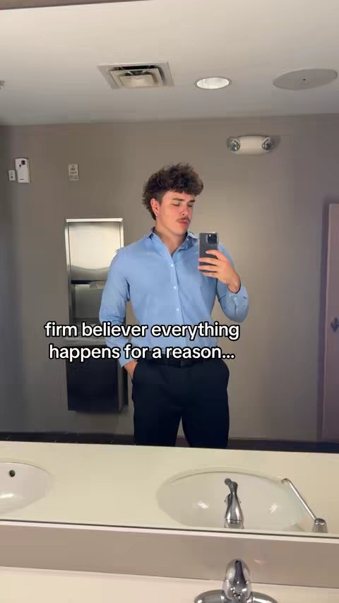A man in a blue shirt and black pants is taking a selfie in a public restroom. The bathroom has a neutral color scheme with a white countertop and a mirror that fills much of the frame. Text on the image reads, "firm believer everything happens for a reason..."